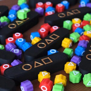 May include: A collection of colorful, geometric game pieces and black rectangular game boards with gold symbols and the letters "OAD". The pieces are red, blue, green, yellow, and purple.