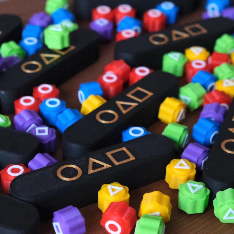 May include: A collection of colorful, geometric game pieces and black rectangular game boards with gold symbols and the letters "OAD". The pieces are red, blue, green, yellow, and purple.