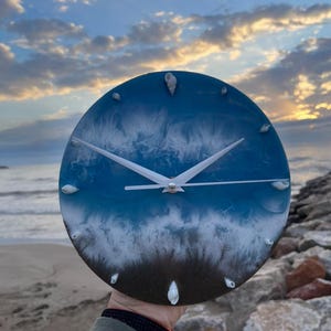 May include: A round resin clock with a blue and white ocean wave design. The clock face has white hands and white seashells around the edge.