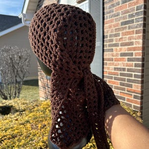 May include: A brown crocheted head wrap with a square pattern. The wrap is tied in a knot at the back of the head and the ends are left to hang down.