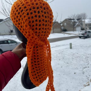 May include: Orange crochet head wrap with long ties. The wrap is made with a simple stitch pattern and has a loose, airy feel.