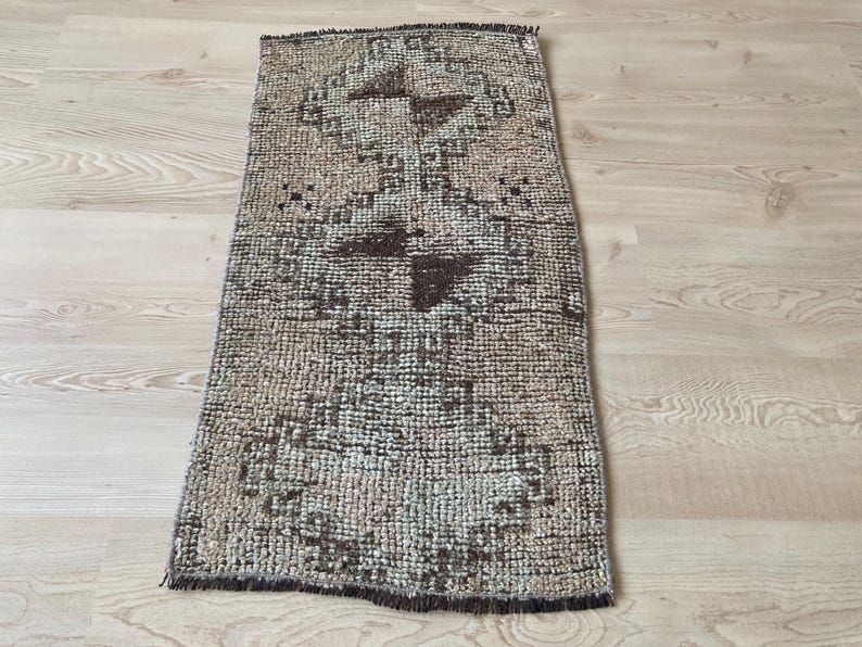 May include: A vintage rug with a geometric pattern in shades of brown and beige. The rug has a fringed edge and is made of a woven material.