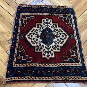 May include: A rectangular area rug featuring a deep red central field with a white and blue geometric medallion pattern. The rug is framed by a dark blue border decorated with small red floral motifs and has short fringe along the top and bottom edges.