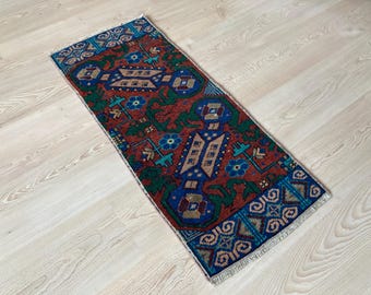 Op de afbeelding: Een rechthoekig vloerkleed met een rode achtergrond en blauwe, groene en bruine geometrische patronen. Het kleed heeft een blauwe rand en franjes aan de korte zijden. Het ligt op een lichtgekleurde houten vloer.