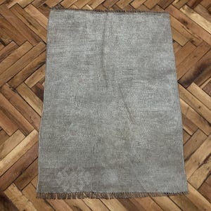 May include: A rectangular, gray rug with a textured surface and a fringed edge. The rug is displayed on a wooden parquet floor. The rug's design features a subtle, all-over pattern.