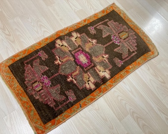 Op de afbeelding: Een rechthoekig vloerkleed met een donkerbruin centraal veld met een geometrisch bloemmotief in tinten magenta, beige en grijs. Het kleed is omlijst door een levendige oranje rand met ingewikkelde, herhalende groene en gele lineaire motieven.