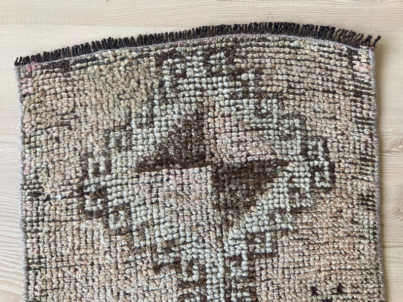 May include: A close-up of a brown and cream patterned rug with a fringe border. The rug has a geometric design with a central cross shape.