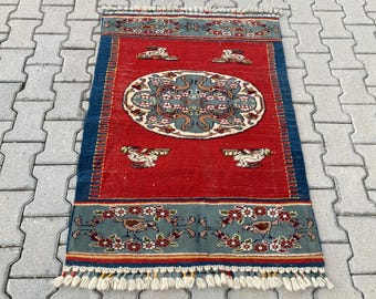May include: A rectangular, hand-woven rug with a red central field. The rug features a central oval design with floral and bird motifs, framed by blue borders with additional bird and floral patterns. The rug has a fringed edge.