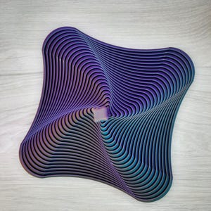 May include: A square-shaped decorative object featuring a series of concentric, layered ridges that create a swirling, wave-like pattern. The object displays a gradient color transition shifting from deep purple to teal and dark blue tones.