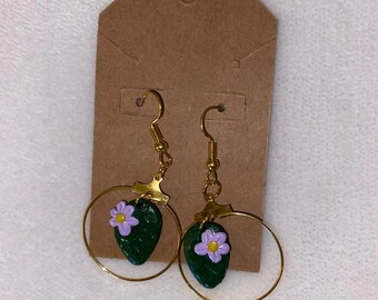 May include: A pair of gold hoop earrings with a green and purple flower charm hanging from each hoop.