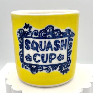 May include: A bright yellow ceramic cup featuring a central white rectangular panel with the dark blue text SQUASH CUP. The text is surrounded by a decorative dark blue floral border, and the cup has a smooth, cylindrical shape with a white rim.