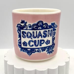 May include: A cylindrical ceramic cup with a light pink exterior and a white interior. The front features a blue hand-painted rectangular frame with floral details, containing the text SQUASH CUP in bold blue capital letters.