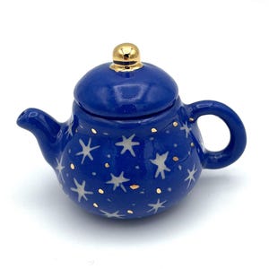 May include: A small, blue ceramic teapot with a lid and handle. The teapot is decorated with white star and gold dot patterns. The lid has a gold knob. The teapot is designed for serving tea.