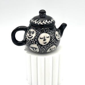 May include: A small, black ceramic teapot with a lid, decorated with white sun-like faces. The handle and spout are black. The lid features a white leaf design. The teapot is displayed on a white, fluted pedestal.