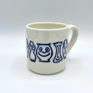 May include: A white ceramic mug featuring a blue line drawing of faces and vases. The mug has a curved handle and a smooth finish. The design is simple and whimsical. The mug is suitable for hot or cold beverages.
