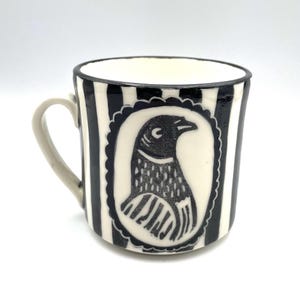 May include: A ceramic mug with a black and white striped design. The mug has a black and white illustration of a bird within a decorative oval frame. The handle is a light grey colour.
