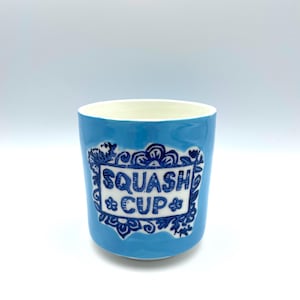 Könnte beinhalten: Eine hellblaue Keramiktasse mit den Worten "SQUASH CUP" in einem weißen dekorativen Rahmen. Die Tasse hat einen weißen Innenraum und eine blaue Außenseite mit floralen und scrollartigen Details.