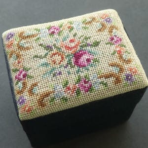 May include: A small, square pincushion featuring a cream-coloured top with a detailed cross-stitch floral pattern in shades of pink, purple, orange, blue, and green. The base of the pincushion is made of a solid black fabric with a soft texture.