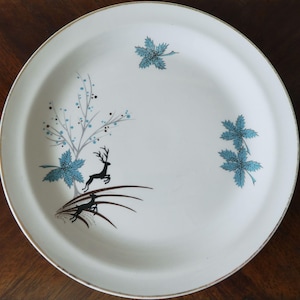 May include: A white ceramic plate with a gold rim, featuring a stylized design of a deer leaping near a tree with blue leaves and branches. Additional blue leaf accents are scattered across the plate's surface. The plate is a vintage piece.