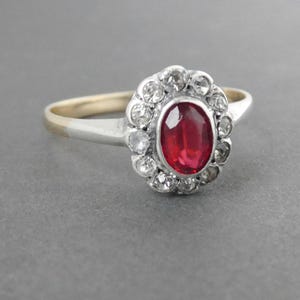 May include: A silver ring featuring a red oval gemstone set in a halo of smaller clear gemstones.