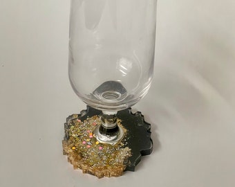 May include: A clear champagne flute sits on a black and gold resin coaster. The coaster has a unique, irregular shape and is embedded with gold and iridescent glitter. The glass is empty and has a long, slender stem.