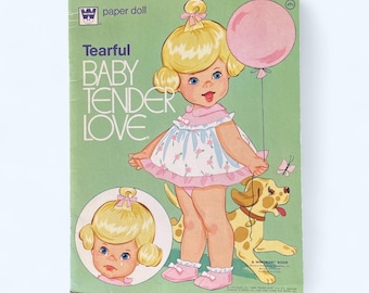 May include: Vintage "Tearful Baby Tender Love" paper doll book. The cover features a blonde-haired doll with a pink balloon and a spotted dog. The doll wears a pink dress with a floral pattern and pink shoes. The background is green.