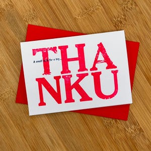 May include: A white card with a red thank you message printed on it. The message reads "THA NKU" with the text "A small thank you for a big..." printed below. The card is sitting on a wooden surface.