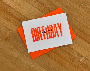 May include: A white card with the words "Happy Birthday" printed in bold, orange letters. The card is folded in half and has an orange envelope.