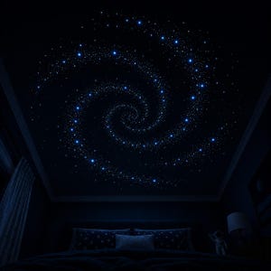 Op de afbeelding: Een donker slaapkamerplafond is versierd met een spiraalvormige melkweg van kleine, helderblauwe sterren. De kamer heeft een bed met kussens met patronen en een nachtlampje, wat een hemelse sfeer cre&euml;ert.