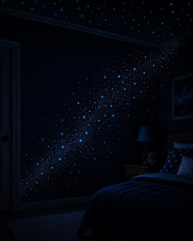 Op de afbeelding: Een donkere slaapkamer met een sterrennachtthema. De muren en het plafond zijn donkerblauw geschilderd, met talrijke kleine, gloeiende witte en blauwe lichten die zijn gerangschikt om op een melkweg te lijken. Een bed met blauw beddengoed en een nachtkastje met een lamp en een teddybeer zijn zichtbaar.