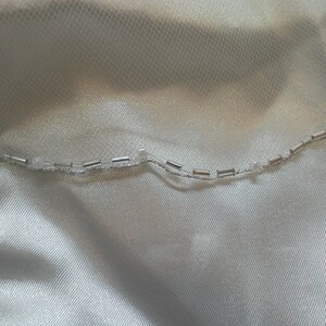 May include: A close-up of a white fabric with a silver beaded trim. The trim is made of small, round beads that are strung together on a thin wire.