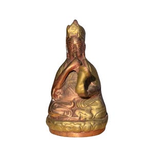 Peut inclure: Statue en bronze d'une figure assise avec une coiffe ornée et détaillée. La statue présente des tons dorés et cuivrés. La figure est dans une pose méditative, une main reposant sur l'autre. La base est également détaillée.