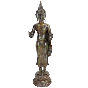 Op de afbeelding: Een staand Boeddhabeeld van brons met een verweerde groene en bruine patina. Het figuur heeft een puntige ushnisha, langwerpige oorlellen en beide handen opgeheven in het Vitarka Mudra-gebaar, op een ronde voet in de vorm van een lotus.