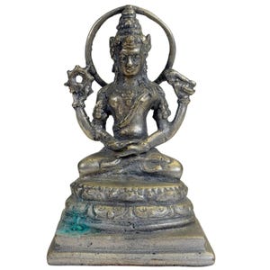 May include: A metallic bronze-toned figurine of a seated deity with four arms, positioned on a tiered rectangular base. The figure features an ornate headdress, a circular halo behind the head, and holds symbolic objects in the upper hands.