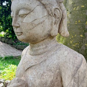 Op de afbeelding: Een verweerd stenen beeld van een figuur in profiel, met een getextureerd oppervlak met zichtbare barsten, een versierde hoofdtooi en een ketting. De figuur heeft gesloten ogen en een serene uitdrukking, met de handen bij de borst.