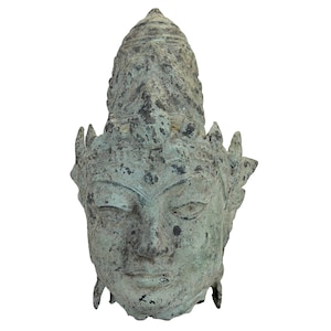 Puede incluir: Una escultura de cabeza en tono bronce desgastado que presenta un tocado alto y puntiagudo con detalles texturizados intrincados. La superficie muestra una pátina moteada con tonos verdes, marrones y gris oscuro, con una expresión serena.