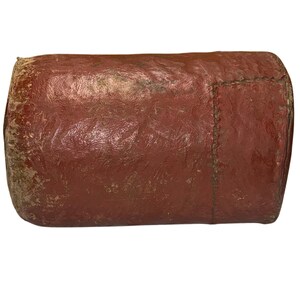 Op de afbeelding: Een cilindrisch, roodbruin leren object met zichtbare slijtage. Het object lijkt aan één uiteinde met de hand gestikt te zijn, wat wijst op een vintage of handgemaakt item. Het leer heeft een textuur, wat een rustieke uitstraling geeft.