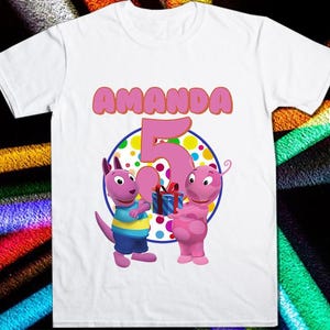 May include: White t-shirt with a pink and orange design featuring two cartoon characters, a kangaroo and a purple creature. The text "AMANDA" and the number "5" are printed in pink on the shirt.