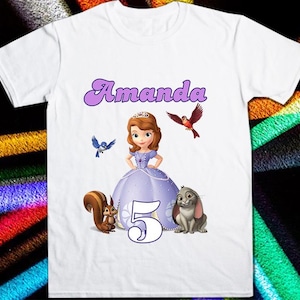 Puede incluir: Camiseta blanca con una princesa de dibujos animados con un vestido morado, con el nombre "Amanda" en escritura morada. El número "5" está debajo de la princesa, con una ardilla y un conejo a cada lado. También aparece un pájaro rojo y un pájaro azul.