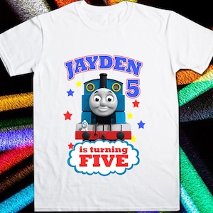 Puede incluir: Camiseta blanca con un diseño de Thomas la locomotora azul y rojo. El texto "Jayden is turning FIVE" está impreso en la camiseta.