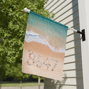 Könnte beinhalten: Eine Strandflagge mit einer Küstenszene. Die Flagge zeigt eine Draufsicht auf einen Sandstrand mit türkisfarbenem Wasser und weißen Wellen. Die Zahl "8647" ist aus Muscheln im Sand geschrieben.