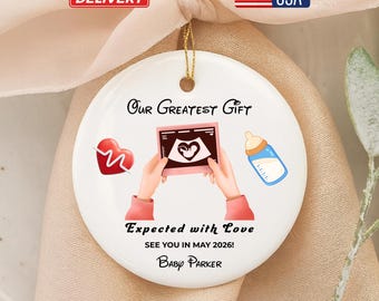 Può includere: Un ornamento in ceramica bianca con un filo d'oro, con la scritta "Our Greatest Gift". Include illustrazioni di un cuore, un'immagine ecografica e un biberon. È presente anche il testo "Expected with Love, See you in May 2026! Baby Parker".