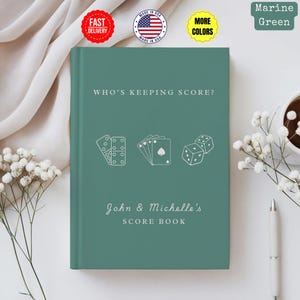 Puede incluir: Un libro de puntuación verde marino con el texto "WHO'S KEEPING SCORE?" y "John & Michelle's SCORE BOOK". La portada presenta ilustraciones de dados, naipes y dominós. El libro está rodeado de flores blancas y un bolígrafo.