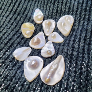 May include: A collection of ten iridescent white and cream-colored mother of pearl shell shapes, arranged on a dark green knitted fabric.