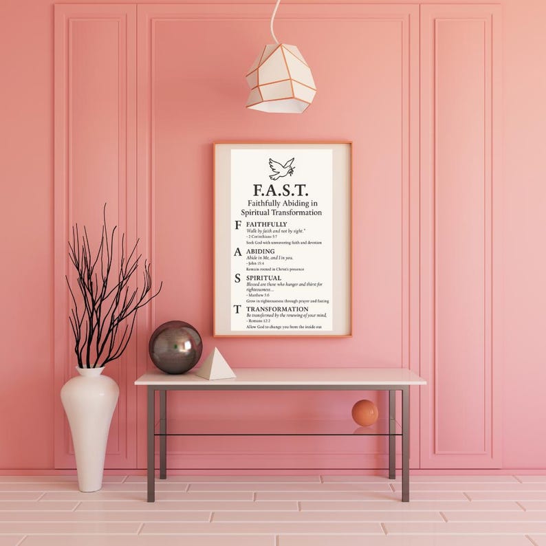 May include: A framed print with the text "F.A.S.T. Faithfully Abiding in Spiritual Transformation" hangs on a pink wall. A white table holds a vase with branches, a silver sphere, and a white geometric shape. A modern pendant light hangs above.