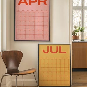 May include: Two framed wall calendars, one pink with "APR" in orange, the other yellow with "JUL" in orange. Both have a grid layout for dates. A wooden chair with metal legs is in the foreground.