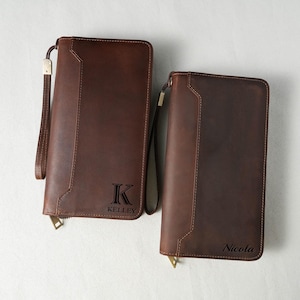 May include: Two brown leather wallets with wrist straps. One wallet has the letter "K" and the name "Kelley" printed on it. The other wallet has the name "Nicola" printed on it. Both wallets have a zipper closure.