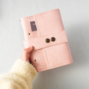 Puede incluir: Un diario de cuero rosa claro con un cierre de solapa asegurado por dos botones de latón antiguo. El diario tiene un diseño cosido y un pequeño bolsillo en la parte delantera. La palabra "LIS" es visible en una tarjeta en el interior.