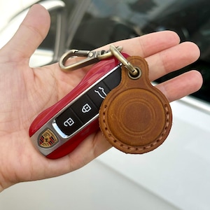 Peut inclure: Clé de voiture rouge avec logo Porsche argenté et boutons noirs. Un porte-clés en cuir marron avec un design circulaire est attaché à la clé. La clé a un fermoir en métal argenté.