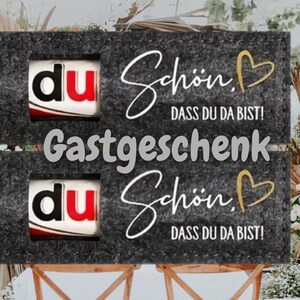 May include: Two dark gray rectangular signs with the text "du", "Sch&ouml;n, DASS DU DA BIST!", and "Gastgeschenk" in white and gold. The signs are placed on top of wooden chairs.